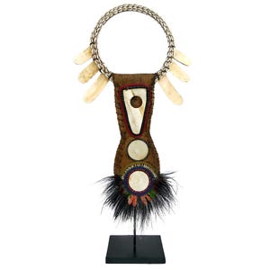 Könnte beinhalten: Eine handgefertigte Halskette mit einem kreisförmigen Muschelkragen und einem zentralen Anhänger. Der Anhänger besteht aus braunem Leder, verziert mit weißen Muschel-Akzenten und einem Büschel schwarzer Federn. Die Halskette wird auf einem schwarzen Ständer präsentiert.