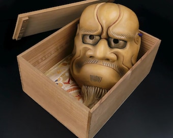 May include: A wooden box with a lid open, revealing a traditional Japanese mask with a fierce expression. The mask is made of wood and painted in shades of brown and yellow. It has a long beard and a stern look.