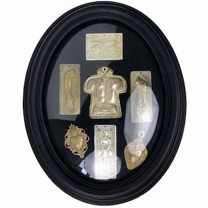 May include: An oval black framed display case with a collection of metal religious charms. The charms include a torso, arms, eyes, a woman, and heart-shaped pendants. The charms are in gold and silver tones.