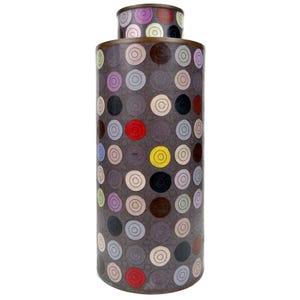May include: A tall, cylindrical vase with a smaller, attached top. The vase is primarily dark gray with a pattern of colorful concentric circles in red, yellow, black, and other hues. The rim and top are a bronze color.