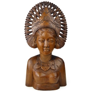 May include: A carved wooden bust of a woman with closed eyes, wearing a detailed headdress and ornate necklace. The sculpture is a warm brown color and appears to be hand-carved, showcasing intricate details and textures. The bust is set against a white background.