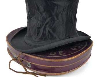May include: A black top hat with a textured, wrinkled crown and a wide brim, resting on a dark purple oval storage box. The box features a brown string closure and the partially visible text DE PARIS printed in white on its lid surface.