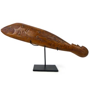 May include: A carved wooden fish sculpture with intricate designs and circular accents. The sculpture is mounted on a black metal stand, showcasing its detailed craftsmanship and unique form. The fish is a warm brown color.