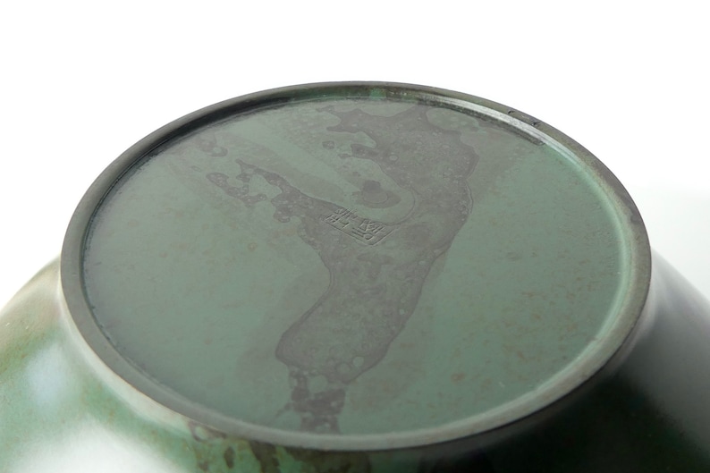 May include: The bottom of a green ceramic bowl with a dark green glaze. The bowl has a mark on the bottom that reads "The New"