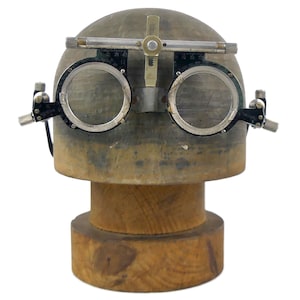 May include: A vintage metal trial frame for optometry, featuring black circular lens holders, adjustable silver bridge components, and side knobs, mounted on a smooth, rounded wooden display head with a cylindrical base for structural support.