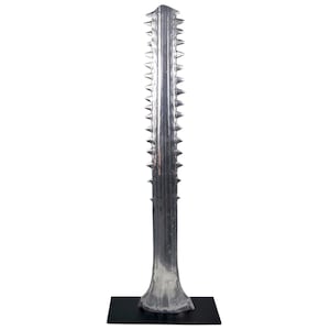 May include: A tall, silver-colored sawfish rostrum sculpture featuring a long, narrow, central blade with a series of sharp, pointed teeth protruding from both sides, mounted vertically on a flat, rectangular black base.