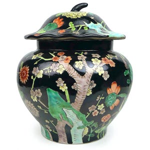 May include: A black ceramic ginger jar with a lid, featuring a hand-painted design of a tree with colorful flowers and birds. The jar has a bulbous shape and a small, black knob on the lid. The overall design is ornate and detailed.
