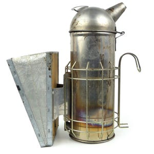 Op de afbeelding: Een vintage metalen bijenroker met een cilindrisch lichaam, een gewelfde bovenkant en een tuit. Het heeft een draadkooi rond het lichaam en een balg aan de zijkant. De roker is gemaakt van zilverkleurig metaal en is ontworpen voor de imkerij.