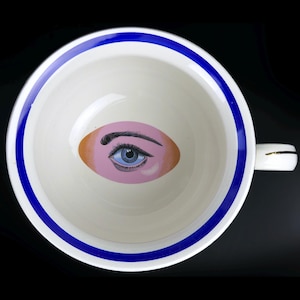 Puede incluir: Un tazón de cerámica blanco con un borde azul y un diseño de ojo pintado en el centro. El ojo es rosa con un iris azul y pestañas negras.