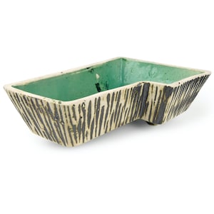 May include: A rectangular ceramic bowl with an irregular, notched side profile. The interior is finished in a smooth, bright mint green glaze, while the exterior features a textured, cream-colored surface with vertical, dark charcoal-colored streaks.