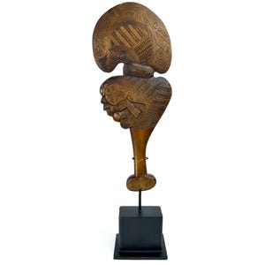 Könnte beinhalten: Eine Holzschnitzerei eines traditionellen Maori Hei Tiki, ein Anhänger, der als Symbol für Glück und Schutz getragen wird. Die Schnitzerei ist aufwendig mit traditionellen Maori-Mustern verziert und auf einem schwarzen Ständer montiert.