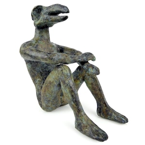 May include: A bronze-toned sculpture of a seated figure with a stylized, elongated head and beak-like features. The figure has a textured, weathered surface with mottled green and brown patina, depicted in a resting pose with arms on knees.