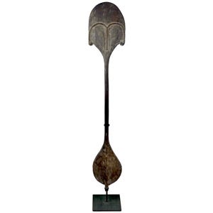 May include: A tall, dark brown wooden paddle featuring a wide, rounded blade at the bottom and a large, flat, semi-circular handle at the top with carved, symmetrical, heart-shaped detailing. The object is mounted vertically on a square metal base.
