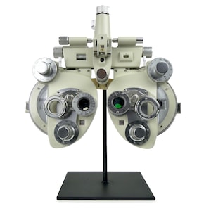 Puede incluir: Un refractor oftalmológico vintage blanco con un soporte de metal negro. El dispositivo tiene varios diales y lentes para exámenes oculares.