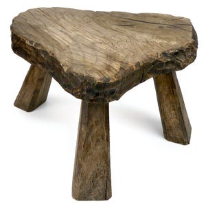 Puede incluir: Un taburete de madera rústico de tres patas con una parte superior irregular y toscamente labrada. El taburete está hecho de madera envejecida, mostrando la veta y la textura naturales. Las patas están anguladas para mayor estabilidad, y el diseño general sugiere un mueble artesanal.