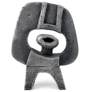May include: A textured, abstract sculpture in shades of grey. The artwork features a rounded head shape with a central oval opening, a cylindrical neck, and a rectangular torso supported by two legs. The sculpture has a rough, industrial aesthetic.