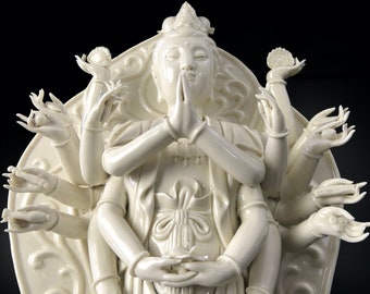 May include: White porcelain sculpture of a multi-armed deity with a serene expression. The deity has a crown and is seated in a lotus position. The sculpture is highly detailed and intricate.