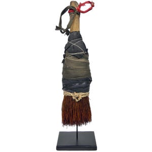 Peut inclure: Un balai fait à la main avec un manche en bois, enveloppé de couches de tissu vert et noir, fixé avec de la ficelle et du fil de fer. Les poils du balai sont brun-rouge. Le balai est exposé sur un support en métal noir.