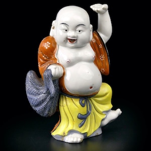 May include: A ceramic figurine of a smiling, bald figure with a large belly. The figure wears a yellow robe, orange shawl, and holds a blue cloth. The figurine is white with painted details and stands on one foot.