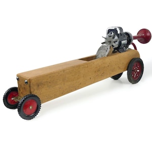 May include: A long, rectangular wooden body mounted on four black wheels with red centres. The vehicle features a metallic motor assembly at the rear, a silver fan-like component, and a prominent red horn extending from the back of the structure.