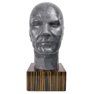 May include: A silver-colored sculpture of a human head with a textured surface. The head is mounted on a wooden base with dark brown and black stripes.