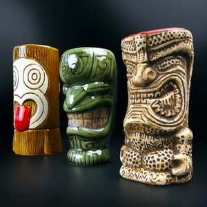 May include: Three tiki mugs with different designs. The first mug is brown with a white face and a red tongue. The second mug is green with a white face and a brown mouth. The third mug is brown with a white face and a brown mouth.