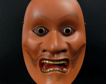 May include: A traditional Japanese Noh mask with a red, orange, and black painted design. The mask has a wide mouth with gold teeth and large, round eyes with black pupils.