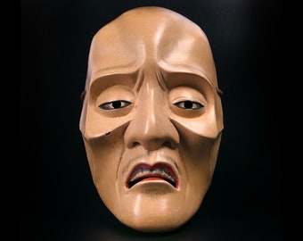 May include: A traditional Japanese Noh mask with a carved wooden face. The mask has a sorrowful expression with closed eyes, a downturned mouth, and a furrowed brow. The mask is painted in shades of brown and beige.