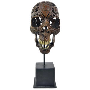 May include: A detailed wooden skull sculpture with carved floral and swirling patterns. The skull features visible teeth and is mounted on a black metal stand with a square base. The sculpture is brown and has an ornate design.