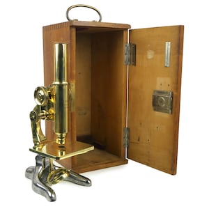 May include: Vintage microscope with brass and silver-colored components, displayed next to its open wooden carrying case. The microscope features a gold-colored tube and a silver-colored base. The wooden case has a handle and metal hinges.