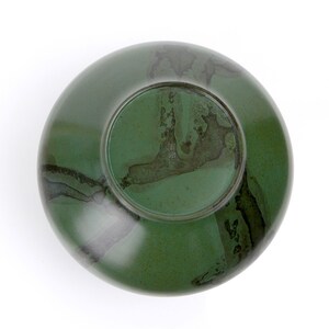 May include: A green ceramic bowl with a dark brown glaze. The bowl has a round shape and a smooth surface. The glaze has a mottled appearance, with darker brown streaks and spots.