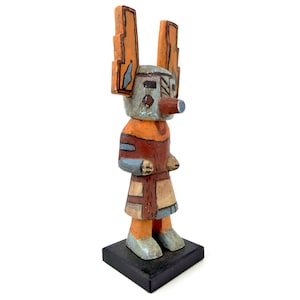 Può includere: Una bambola kachina in legno intagliato a mano su una base quadrata nera. La figura presenta un volto azzurro con un muso prominente, alte orecchie arancioni e un corpo dipinto in tonalità arancione, rosso, beige e blu con motivi geometrici.