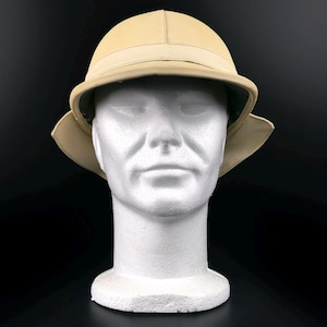 Può includere: Un elmetto coloniale beige con tesa larga e una fascia colorata chiara attorno alla corona. Il cappello è esposto su una testa di manichino bianca su sfondo nero. L'interno del cappello è verde scuro.