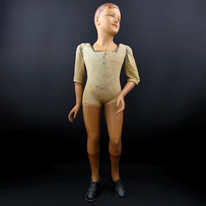 Könnte beinhalten: Eine Vintage-Kinderschaufensterpuppe mit bemaltem Gesicht und modelliertem Haar, bekleidet mit einem beigen, kurzärmeligen Oberteil mit Schnürung und sichtbarem Textaufdruck. Die Puppe hat bewegliche Arme und trägt dunkle Schnürschuhe.