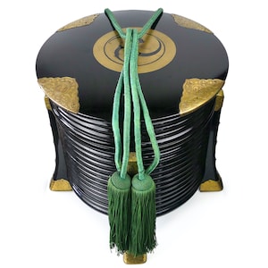 May include: A black lacquered box with gold accents and a green tassel. The box is made of wood and has a round shape. It is decorated with a gold design on the top.