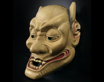 May include: A traditional Japanese wooden mask with a fierce expression. The mask is painted in shades of yellow, red, and black, with detailed carvings and painted lines. The mask has large, exaggerated eyes and a wide, open mouth with sharp teeth.