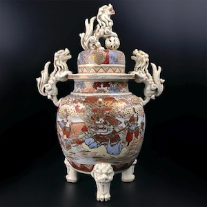 Puede incluir: Un jarrón de cerámica japonesa antiguo y detallado con tapa, que presenta una escena pintada colorida de guerreros. El jarrón tiene tres patas con forma de león y dos asas de león. Una estatua de león se encuentra encima de la tapa. El jarrón es principalmente crema, rojo, azul y dorado.