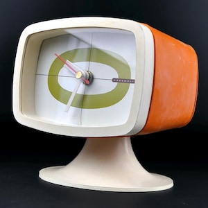 Könnte beinhalten: Eine fernsehförmige Analog-Uhr mit orangefarbenem Außengehäuse und weißem Standfuß. Das Zifferblatt hat einen weißen Hintergrund mit einem großen olivgrünen Oval, dünnen schwarzen Fadenkreuzen und dem Wort ASSEMBLY in einem kleinen Rechteck.
