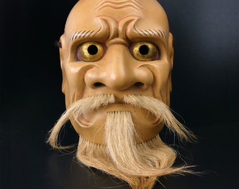 May include: A traditional Japanese wooden mask with a carved face, painted in shades of brown and gold. The mask has a long white beard and mustache made of real hair. The eyes are large and round with gold accents.
