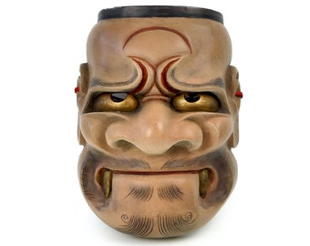 May include: A traditional Japanese wooden mask with a fierce expression. The mask is painted in shades of brown and gold, with red accents. The eyes are large and round, and the mouth is open in a wide grin, revealing sharp teeth. The mask has a detailed beard and mustache.