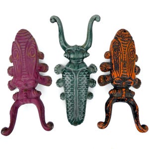 Może przedstawiać: Trzy dekoracyjne metalowe rzeźby owadów w odcieniach fioletu, turkusu i pomarańczy. Każdy owad ma unikalny design ze skomplikowanymi detalami i jest zamontowany na trójnożnej podstawie. Rzeźby są eksponowane na białym tle.