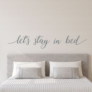 May include: A white bed with a gray headboard and a white comforter. The wall behind the bed has a gray vinyl decal that reads "let's stay in bed".
