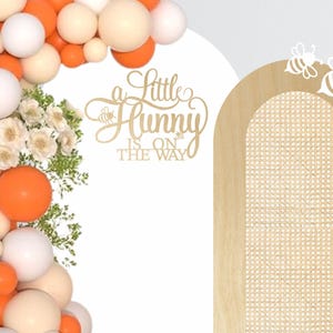 May include: A decorative baby shower backdrop with a balloon arch in orange, white, and cream colors. The arch is next to a wooden arch with a woven pattern and bee cutouts. The text "A Little Hunny Is On The Way" is displayed.