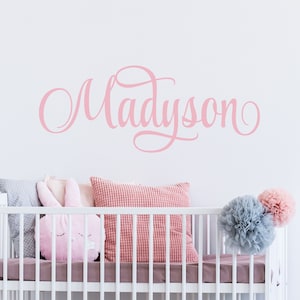 May include: A white crib with a pink and gray patterned pillow and a pink bunny pillow. The wall behind the crib has a pink cursive name "Madyson" written on it.