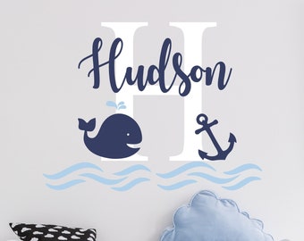 May include: A white letter H with the name "Hudson" written in blue cursive above it. A blue whale with a water spout is to the left of the letter and a blue anchor is to the right. The letter, whale, and anchor are all on top of blue wavy lines that represent water.