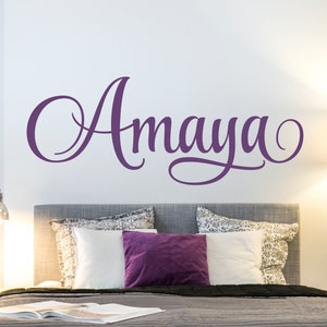 May include: Purple vinyl wall decal with the name "Amaya" written in a cursive font. The decal is on a white wall above a gray headboard with pillows.