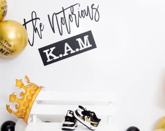 Könnte beinhalten: Eine weiße Holzbank mit einem Paar schwarz-goldenen Sneakers darauf. Im Hintergrund befinden sich goldene und schwarze Ballons, sowie eine goldene Krone und ein Schild mit der Aufschrift "The Notorious K.A.M."