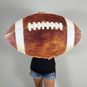 May include: A large, brown and white football prop with detailed stitching and stripes. The football is held in front of a person, obscuring their upper body. The prop is designed to resemble a real football.