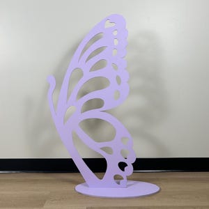 May include: A large, lavender-colored butterfly cutout, standing on a round base. The butterfly has intricate details and a delicate design.