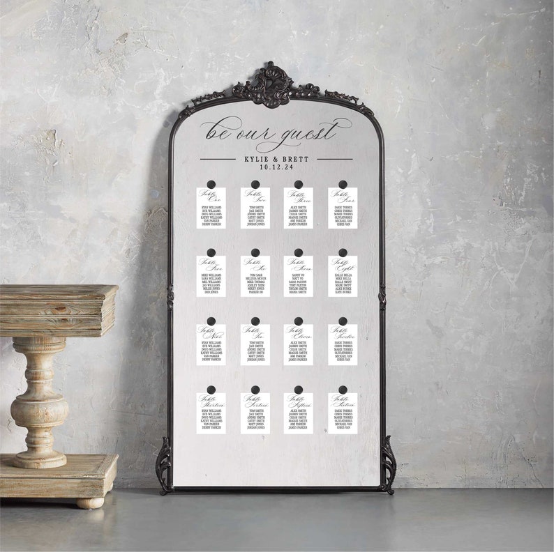 May include: A black framed mirror with a white background and the text "be our guest" in cursive script. Below the text is "KYLIE & BRETT 10.12.24". The mirror is decorated with small white cards with black text, each labeled "Table" followed by a number. The cards are attached to the mirror with black dots.